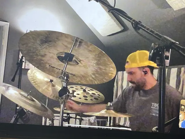 Drummer in a yellow cap playing the house kit, seen through the cymbals