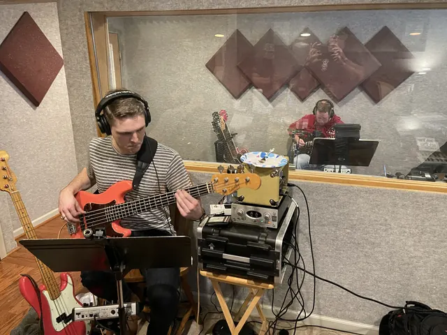 Bass player in headphones tracking in the live room, with a guitarist behind the glass