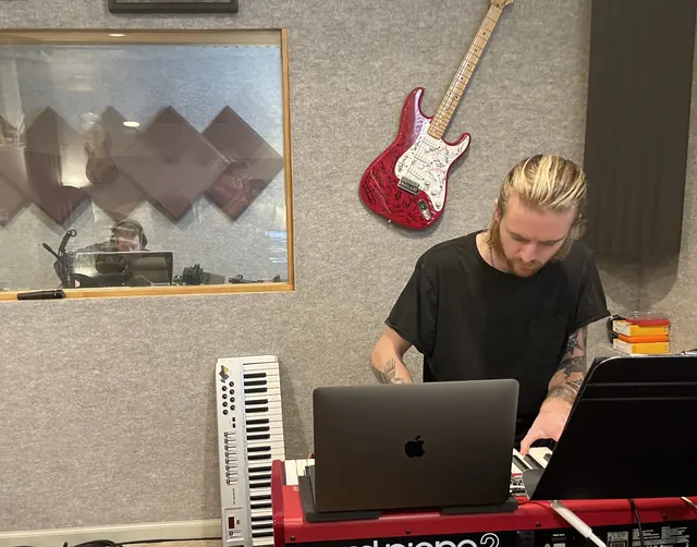 Keyboard player working at a laptop on a red Nord, with the control room window behind