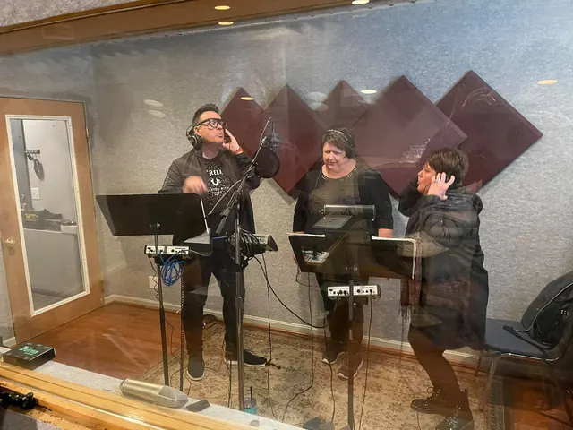 Wide view of three singers recording in the vocal booth through the glass