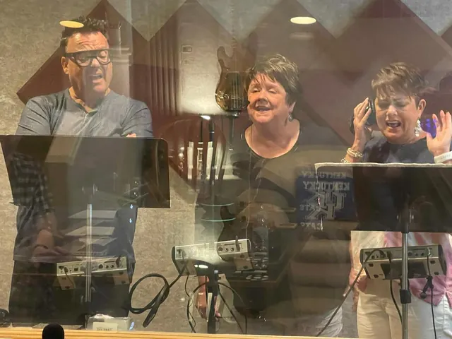 Three singers at microphones in the vocal booth, seen through the control room glass